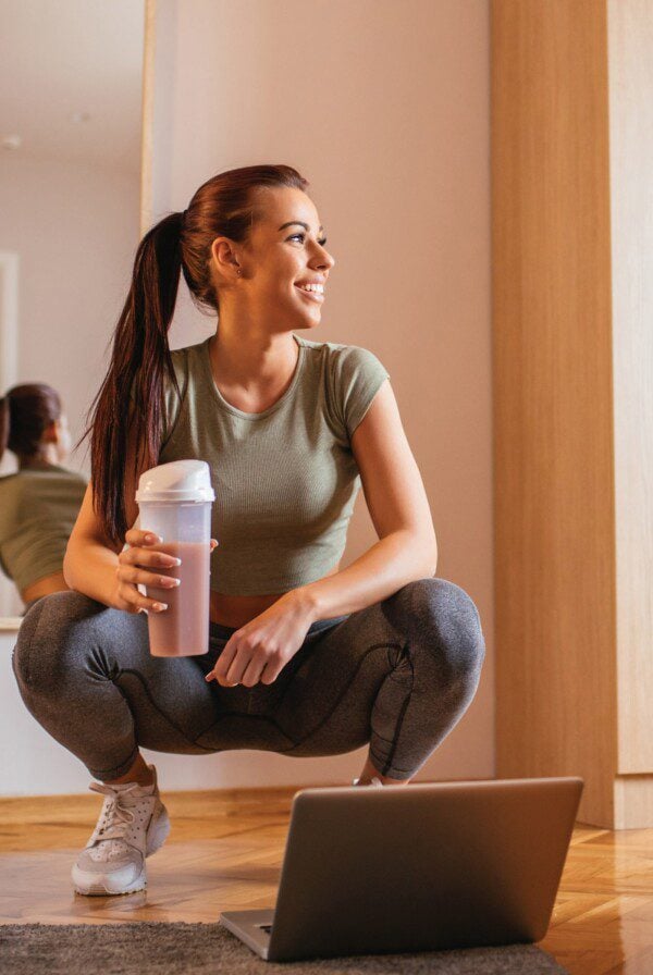 Fit woman with a shaker cup crouched over a laptop.