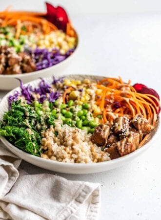 Brown rice, edamame, tempeh, carrot, corn, kale and red cabbage in a bowl topped with sauce and sesame seeds.