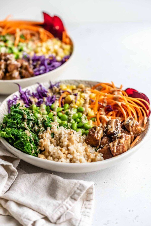 Brown rice, edamame, tempeh, carrot, corn, kale and red cabbage in a bowl topped with sauce and sesame seeds.