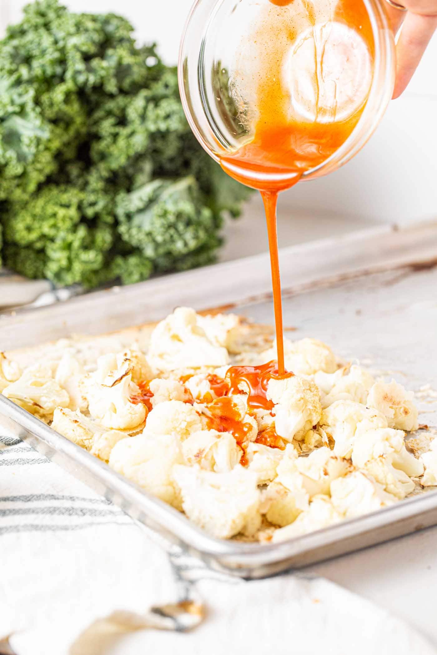 Crispy Buffalo Cauliflower Kale Salad 5 A baking pan filled with golden-brown roasted cauliflower, being drizzled generously with spicy buffalo sauce, ready for its final bake.