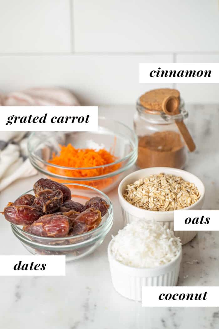 Oats, dates, grated carrot and dried coconut in bowls on a counter.