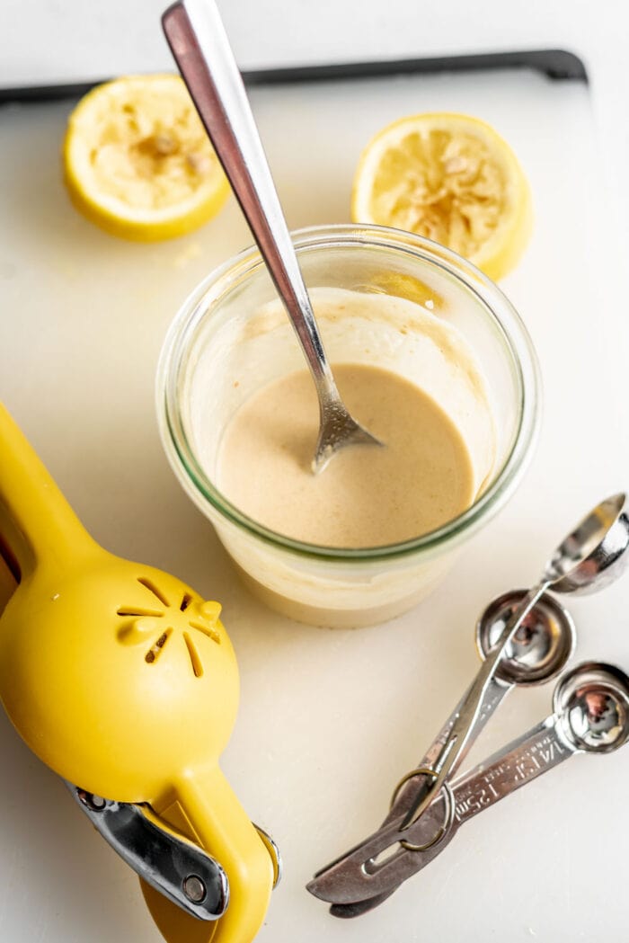 Tahini dressing in a glass jar with a spoon sitting on a cutting board beside lemons.