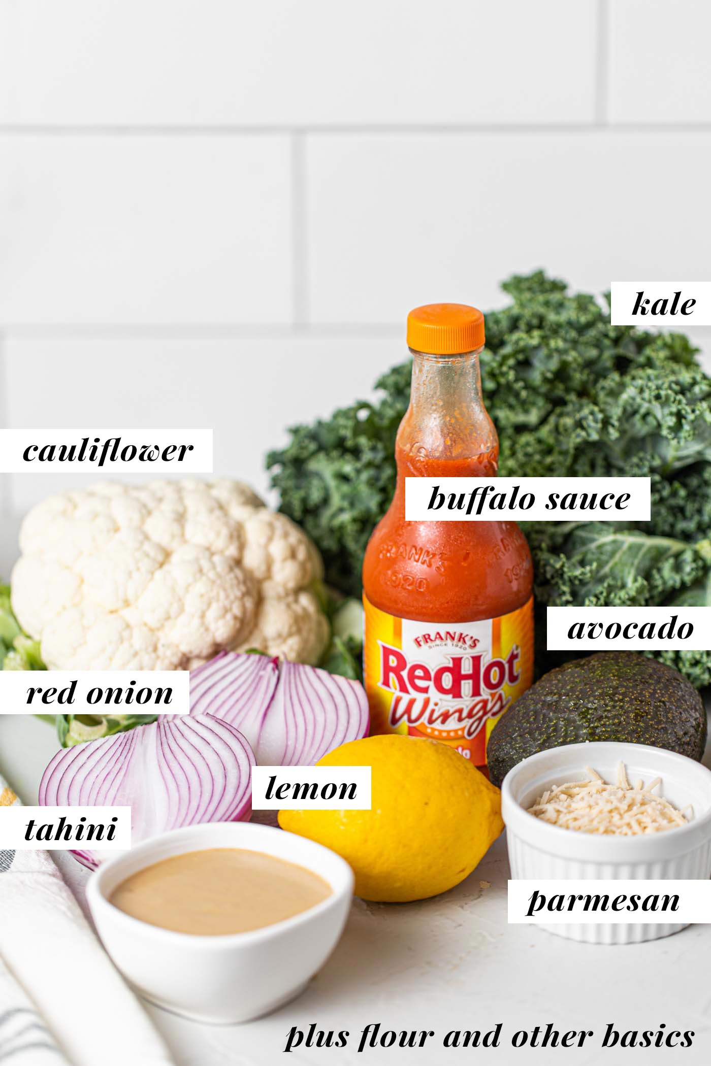 Crispy Buffalo Cauliflower Kale Salad 3 A collection of fresh ingredients laid out on a kitchen counter, including a head of cauliflower, vibrant kale, ripe avocado, lemons, a bottle of hot sauce, and a jar of tahini, ready for meal preparation.