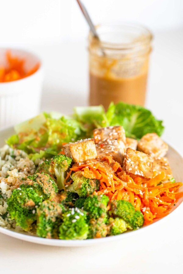 A bowl with carrot, tofu, broccoli, cauliflower rice, tofu and peanut sauce.