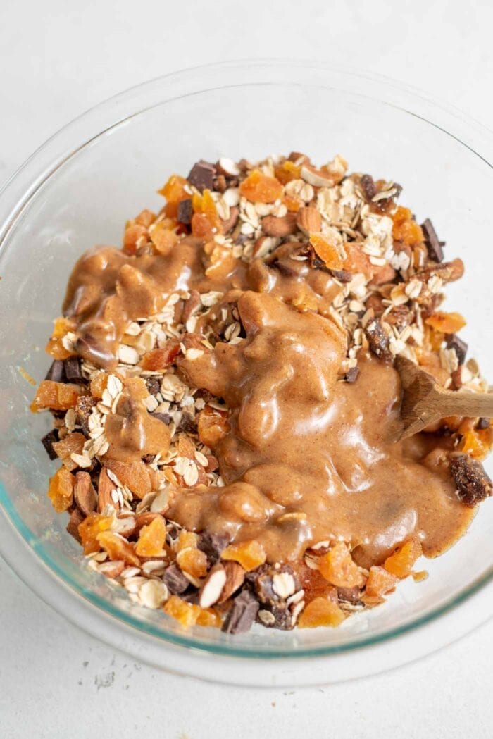Almonds, oats, dates, apricots and chocolate with maple syrup in a glass mixing bowl.