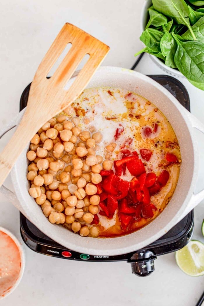 Diced tomato, rice, coconut milk and chickpeas cooking in a pot with a wooden spoon.