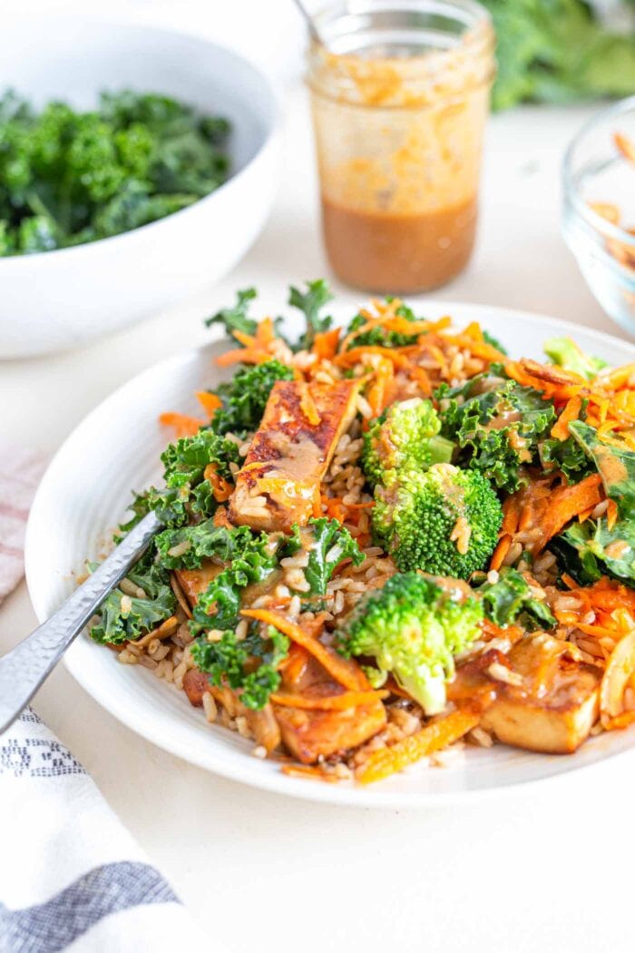 A bowl with shredded carrot, broccoli, tofu, kale and a creamy sauce.