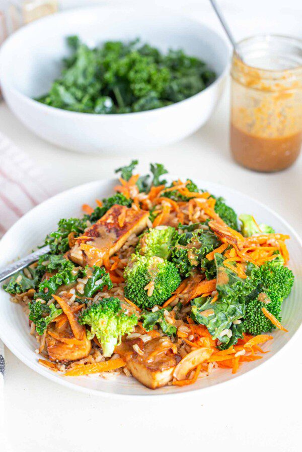 A bowl with shredded carrot, broccoli, tofu, kale and a creamy sauce.