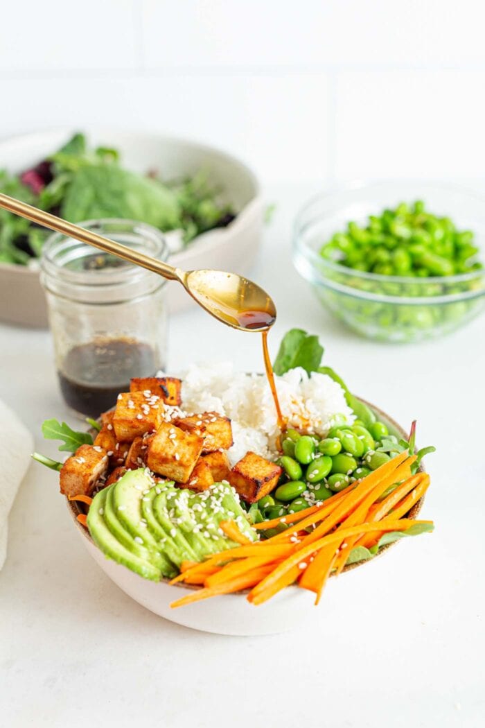 Drizzling a spoonful of sauce over a bowl of greens, avocado, carrot and tofu.