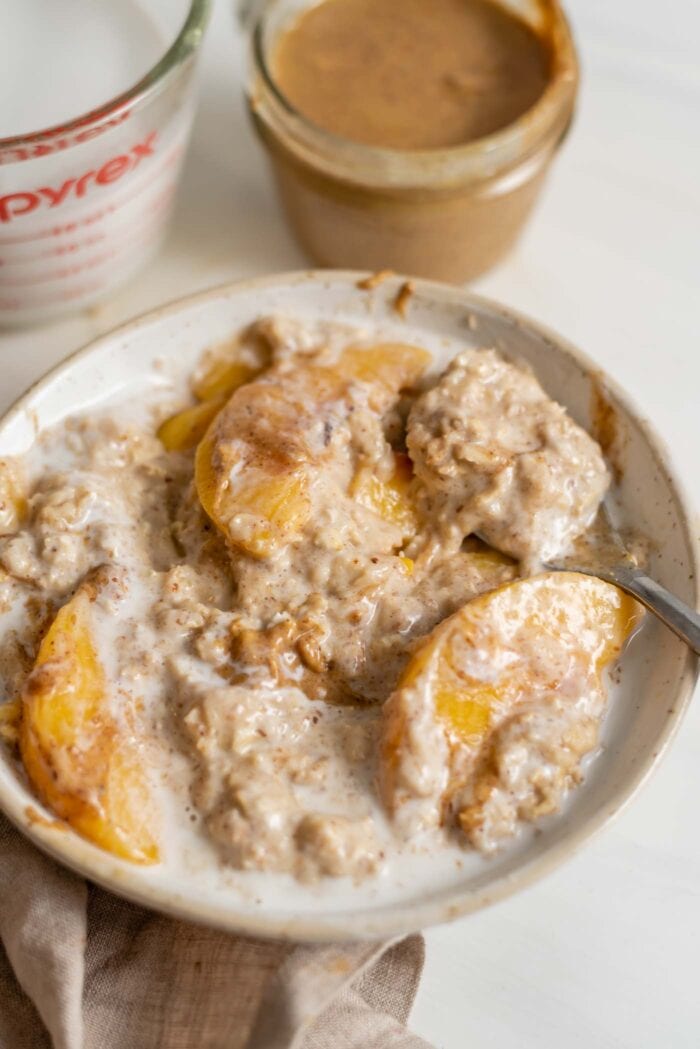 A bowl of peaches and cream oatmeal with a spoon in it. A container of almond butter and coconut milk are in the background.
