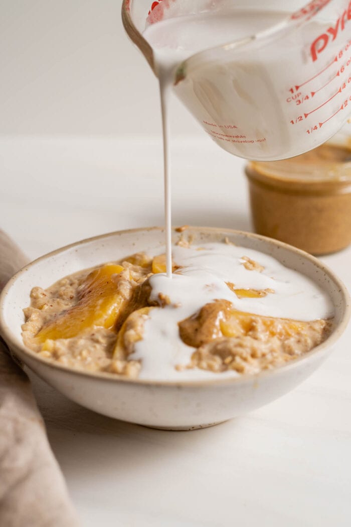 A measuring cup pouring milk into a bowl of peach oatmeal.