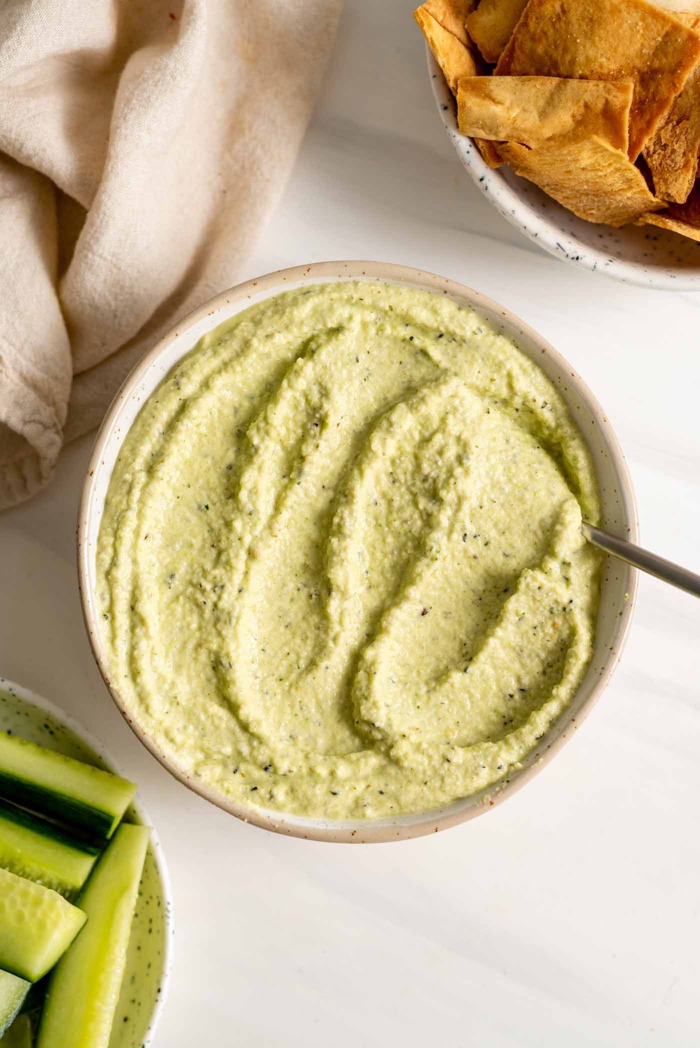 A bowl of green hummus with some chopped cucumber and pita chips.