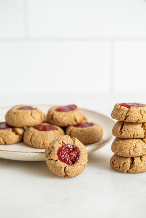 A jam-filled small thumbprint cookie leaning against a plate of cookies with a stack of 4 cookies beside it.