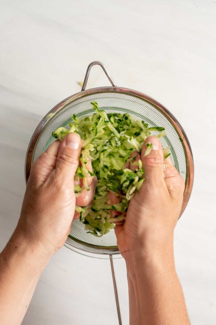Two hands squeezing grated cucumber over a colander.