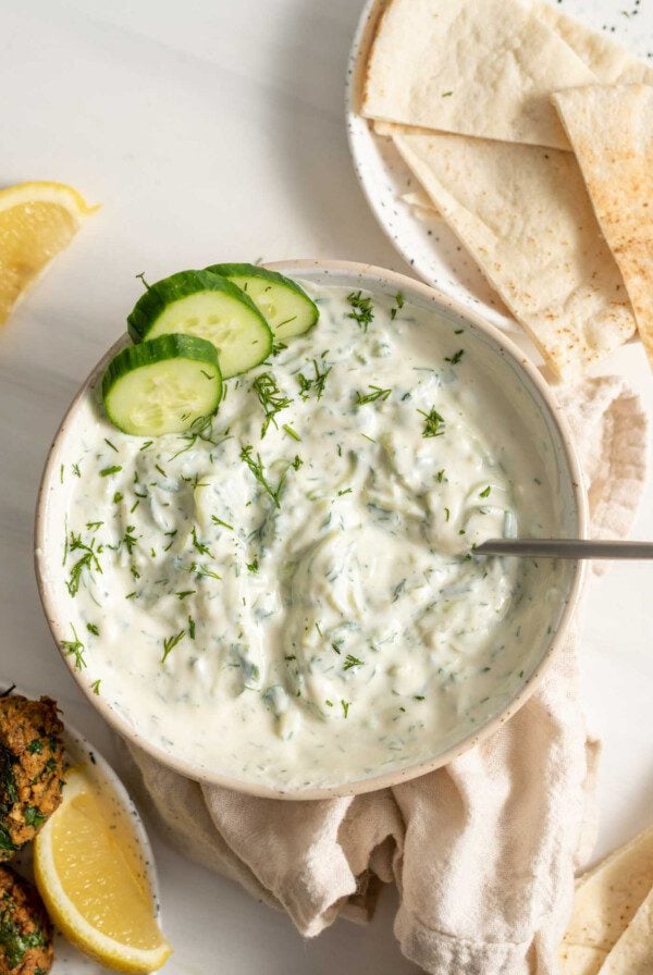 A bowl of tzatziki dip with some pita on a plate beside it.