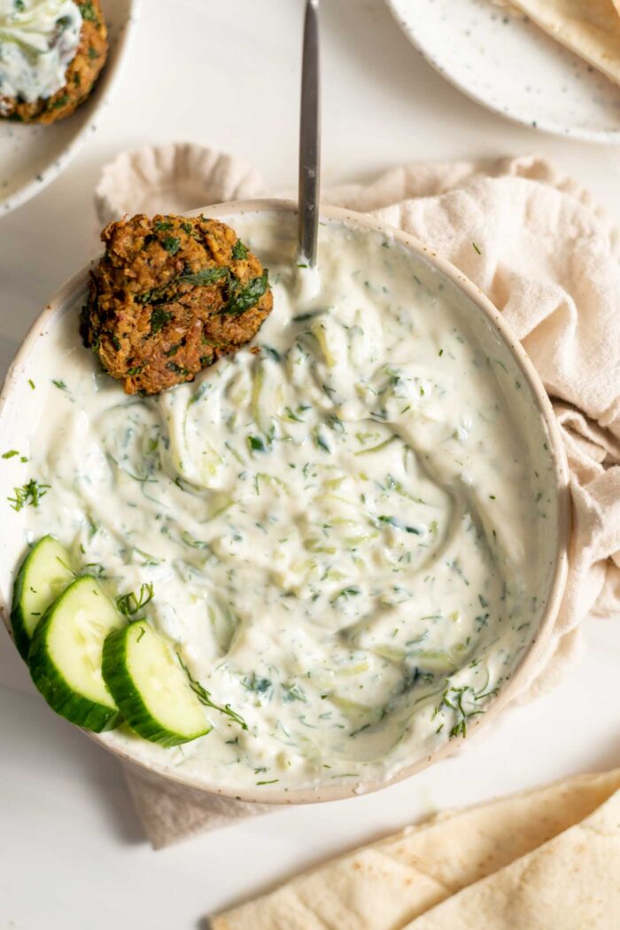 A bowl of tzatziki with a falafel and a few slices of cucumber in it.