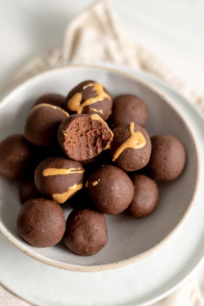 Chocolate energy balls drizzled with peanut butter in a bowl, one with a bite taken out of it to show inside.