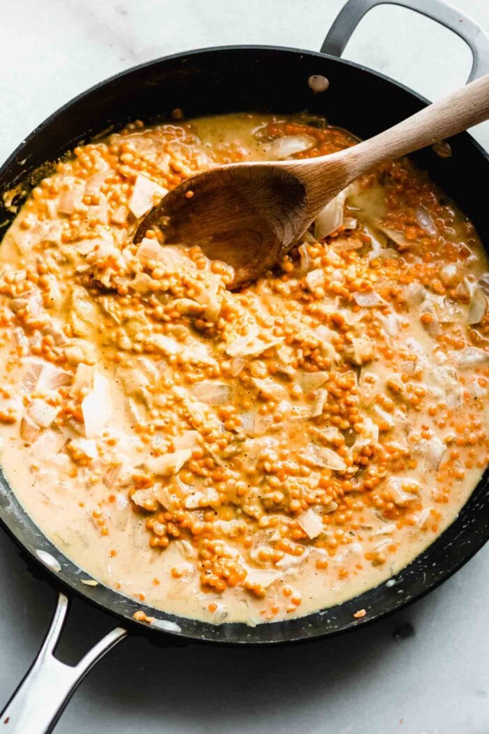 A creamy red lentil and coconut milk mixture in a skillet with a wooden spoon.