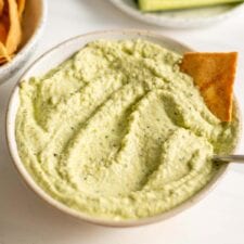 A bowl of green hummus with some chopped cucumber and pita chips.