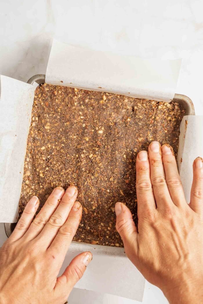 Two hands flattening energy bar dough in a a parchment paper-lined metal baking pan.