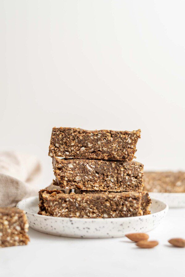 3 raw energy bars stacked on a small round plate with some almonds in the foreground.