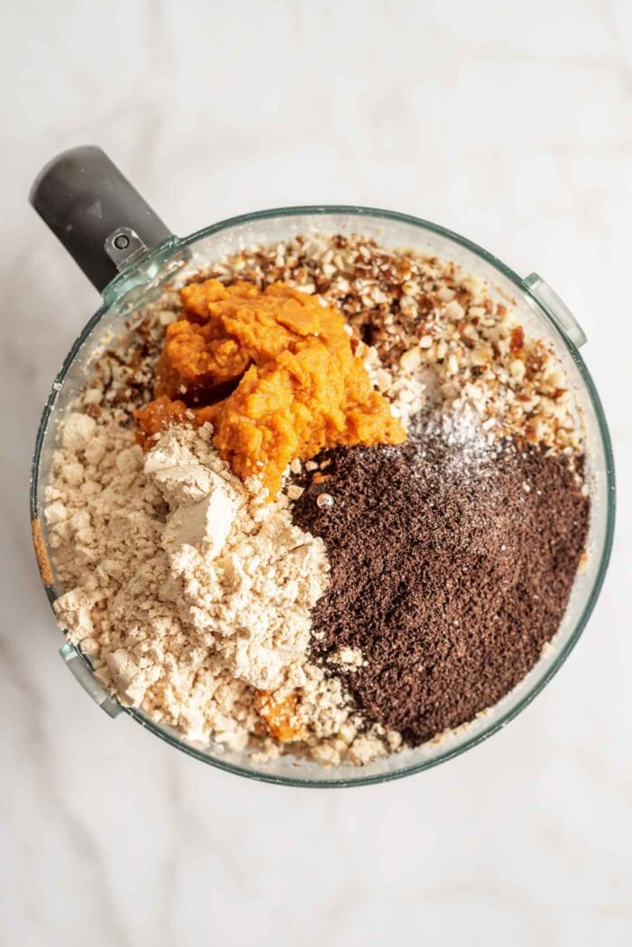 Pumpkin puree, ground coffee and protein powder sitting on top of a crumbly mixture in a food processor.