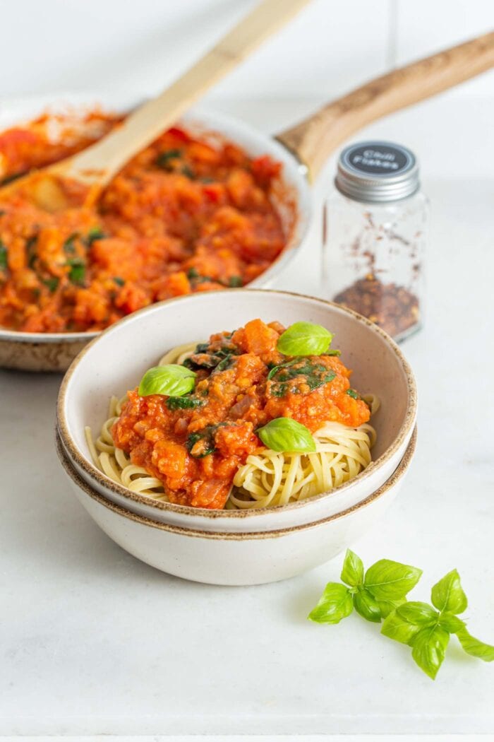 A bowl of tomato pasta with fresh basil, skillet of tomato sauce in the backgroun.