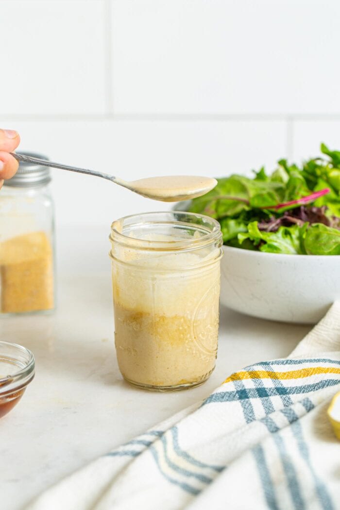 Spooning a creamy tahini dressing out a jar. Bowl of green salad in background.