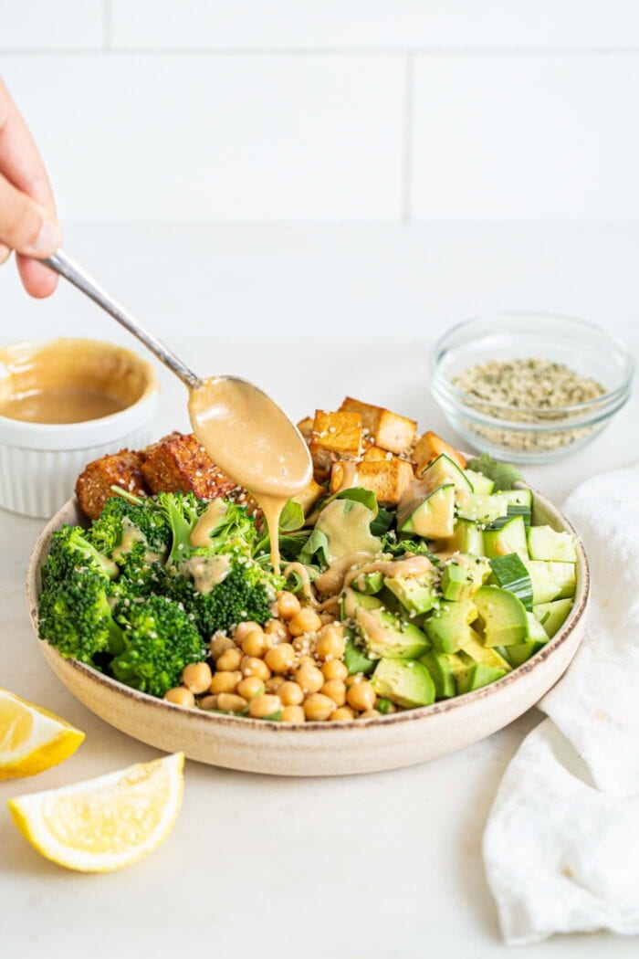 Adding a spoonful of tahini to a bowl of chickpea, broccoli, tofu, cucumber and tempeh salad.