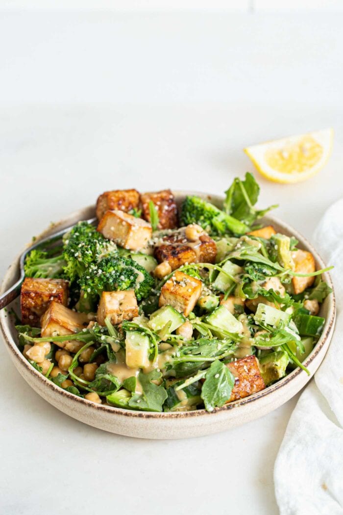 A bowl of salad with arugula, avocado, cucumber, tofu and tempeh. Slice of lemon in background.
