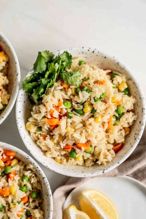 A bowl of rice pilaf with peas, corn and carrots and some cilantro on the side, two more bowls sitting just out of the frame.