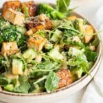 A bowl of salad with arugula, avocado, cucumber, tofu and tempeh. Slice of lemon in background.