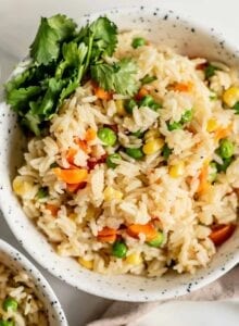 A bowl of rice pilaf with peas, corn and carrots and some cilantro on the side.