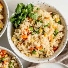 A bowl of rice pilaf with peas, corn and carrots and some cilantro on the side.