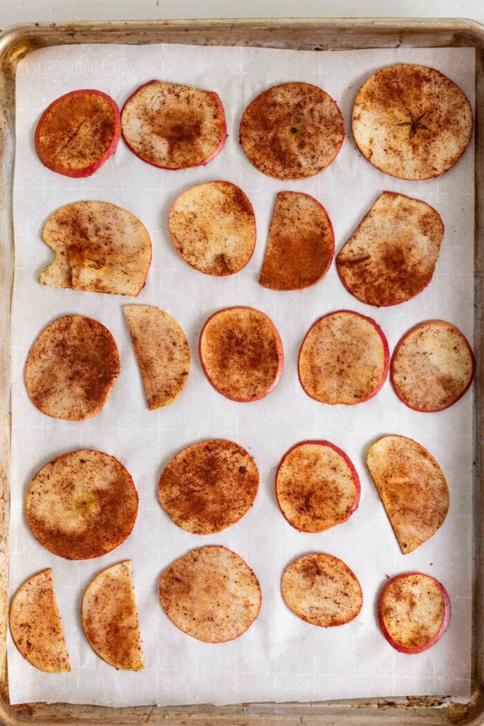 Thin slices of apple coated in cinnamon spread on a parchment paper-lined baking tray.