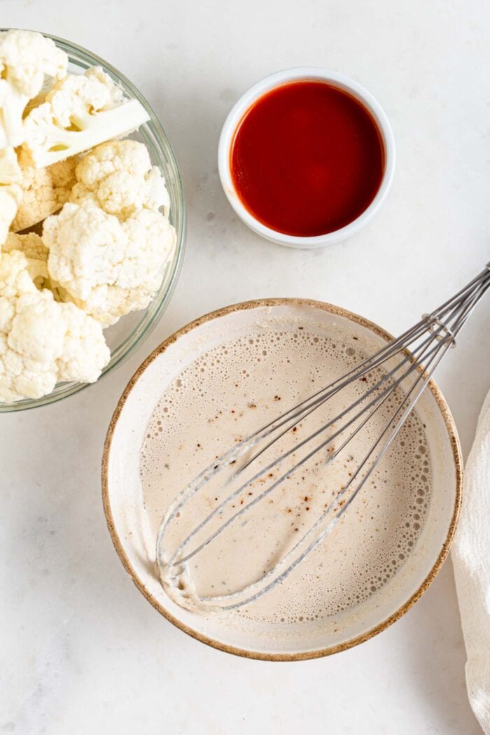 A bowl of flour-based batter with a whisk in it, bowl of cauliflower beside it.