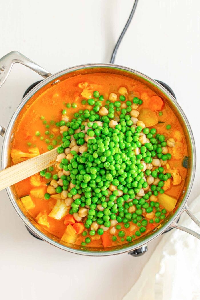 Adding peas and chickpeas to a large pot of stew. Wooden spoon rests in pot.