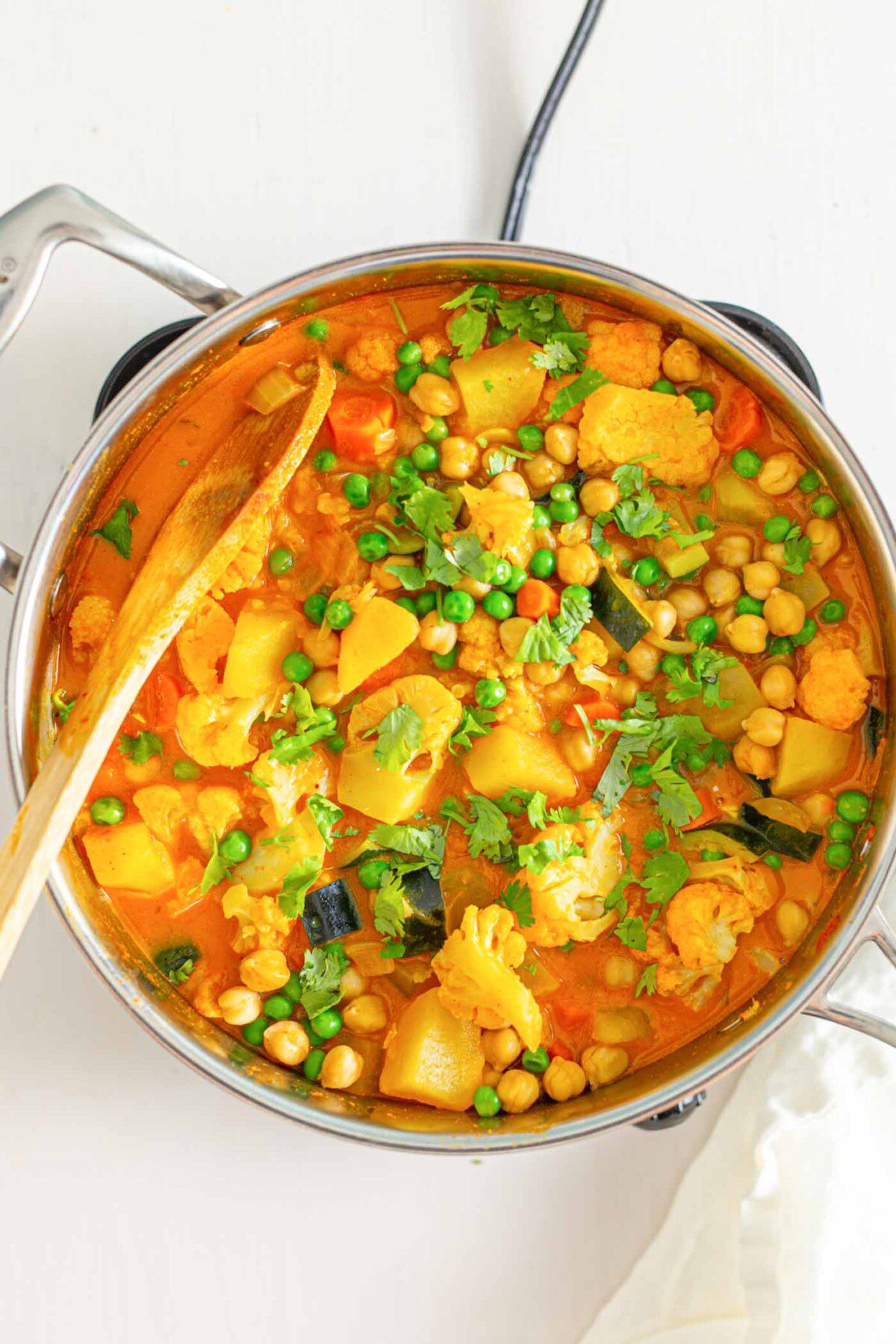 Pot of chickpea and potato curry with peas and a wooden spoon in the pot.