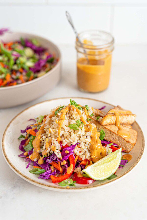 Bowl of colorful salad with brown rice, peanut sauce, tofu and veggies. Jar of sauce in background.