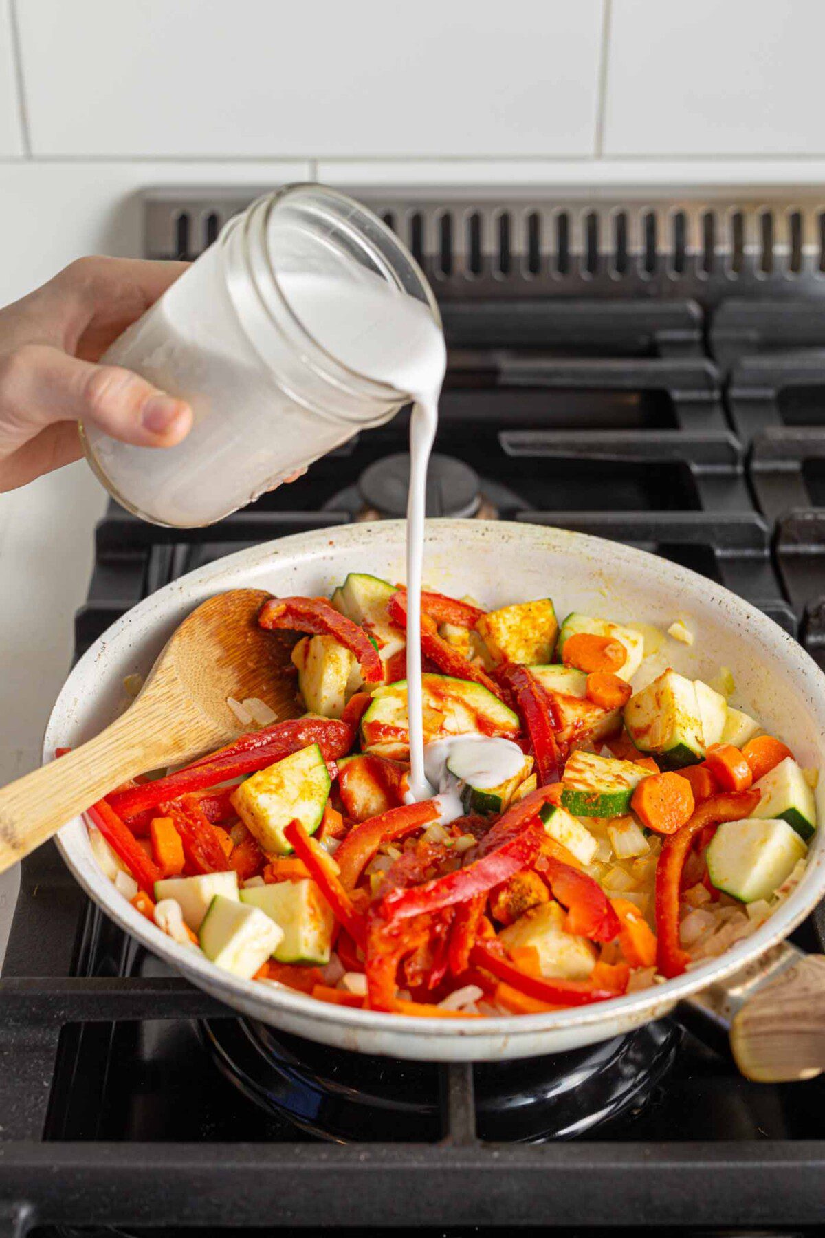 Pouring coconut milk into a vegetable stir fry cooking on a stovetop.