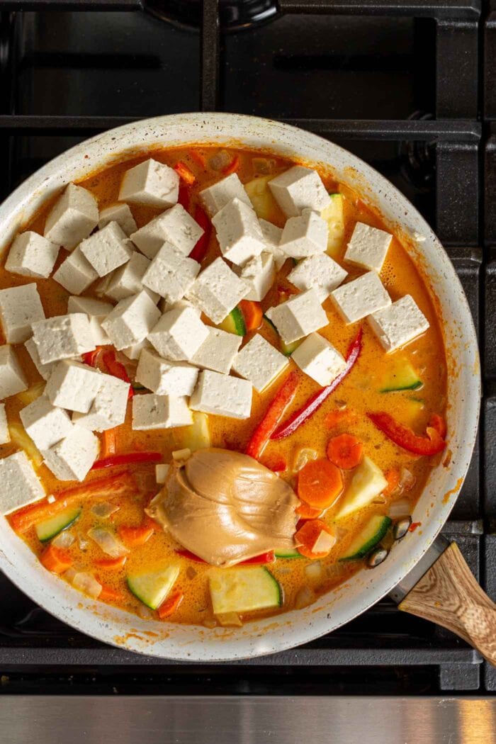 Tofu and peanut butter being added to a vegetable curry in a skillet.