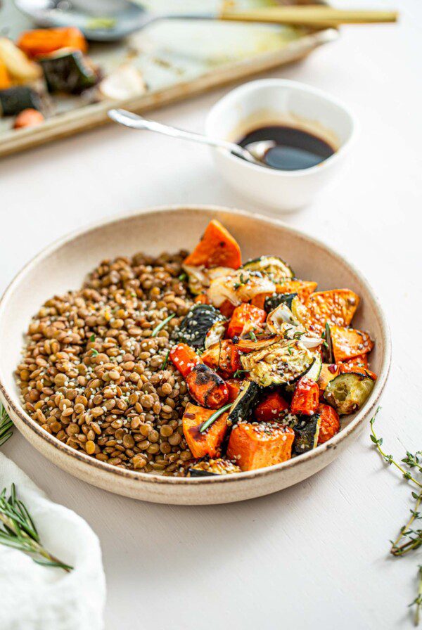 Lentils and roasted vegetables in a bowl. Small dish of balsamic dressing in background.