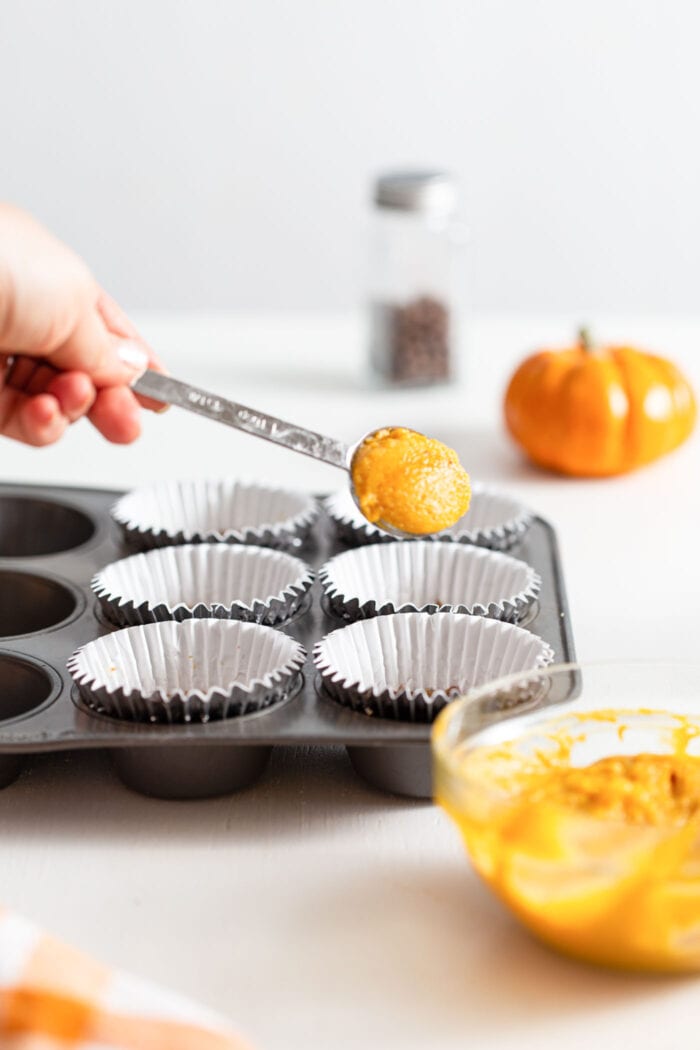 Hand adding a spoonful of pumpkin puree to a lined muffin tin. There is a small pumpkin in the background and a bowl of pumpkin puree in the foreground.