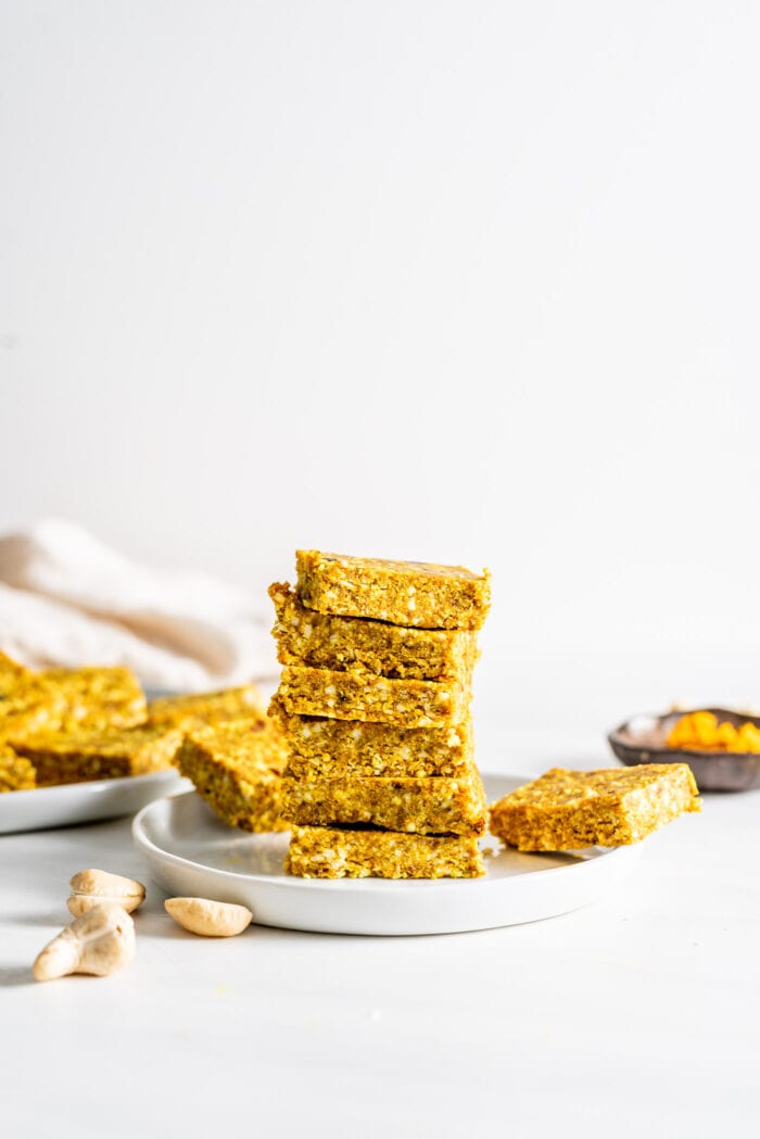 Stack of 6 turmeric coconut and cashew energy bars on a small plate. More bars in background. Cashews scattered about.