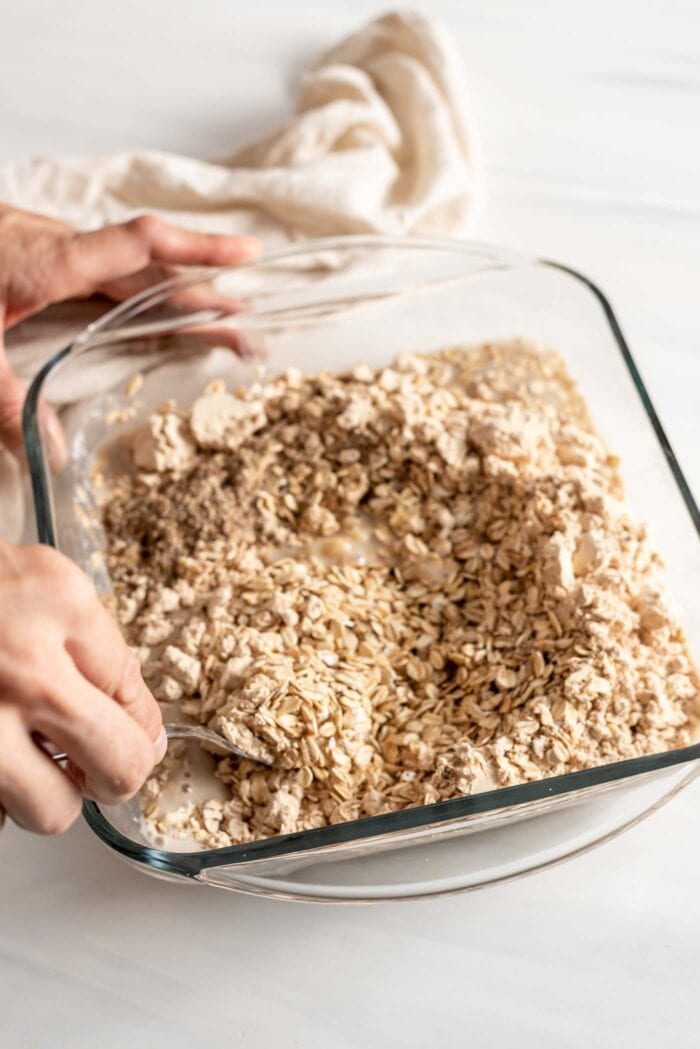 Two hands mixing together oats and protein powder in a glass baking dish.