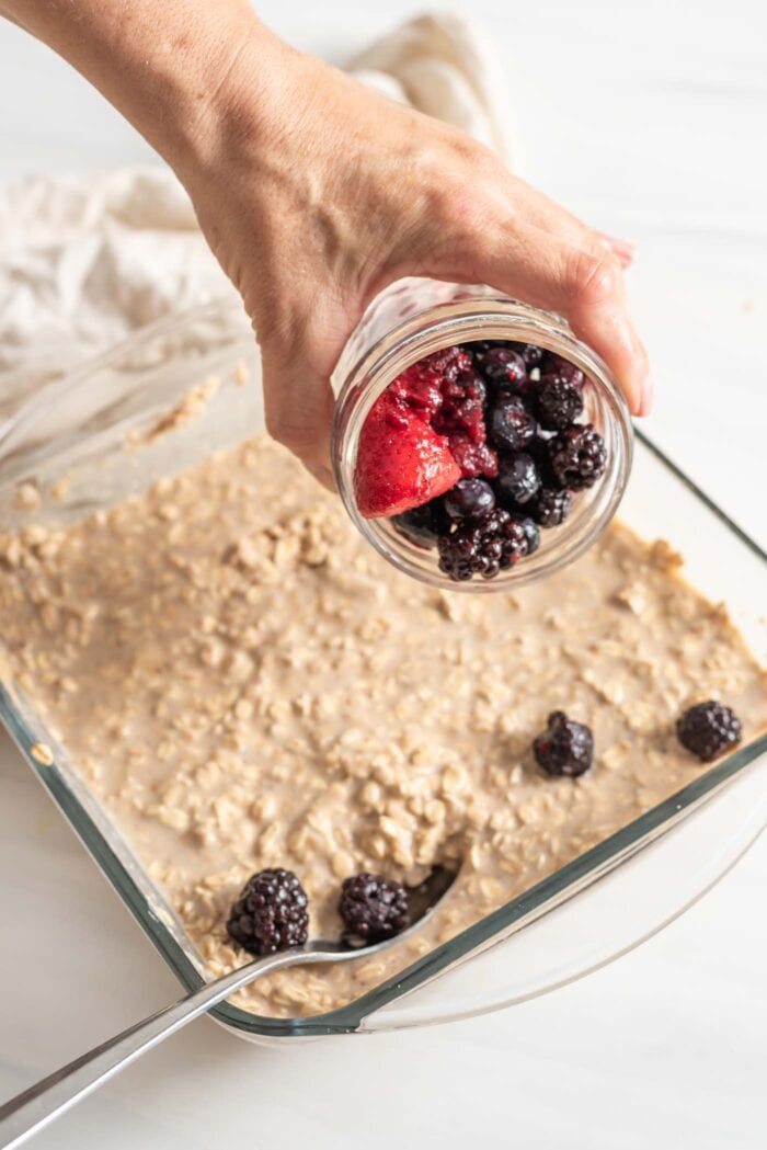 Hand sprinkling a jar of frozen berries over a baking dish of oatmeal.