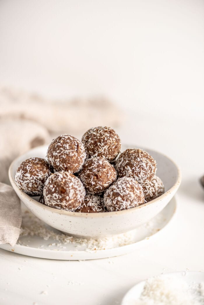 Bowl of protein balls rolled in coconut. Bowl sits on a plate, dish cloth in background.
