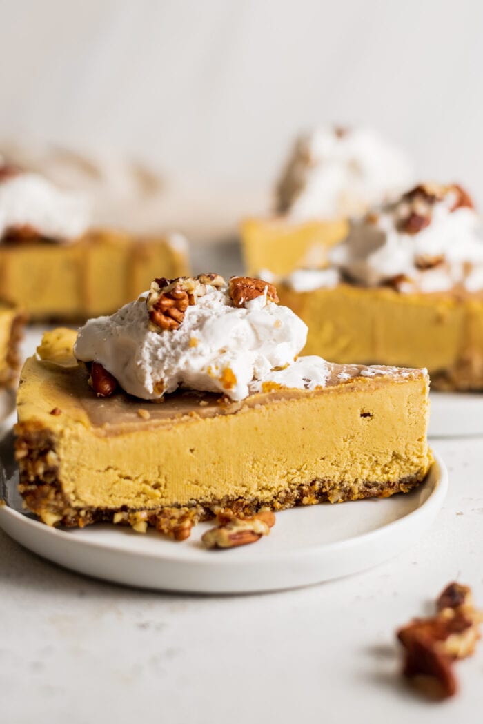 A slice of vegan no-bake pumpkin cheesecake topped with whipped cream and pecans on a plate. There are more slices of pie on plates in the background.
