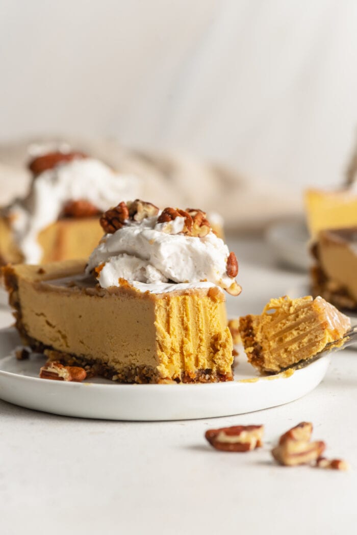Slice of pumpkin cheesecake topped with whipped cream on a small plate. Bite taken out of slice of cake with a fork.