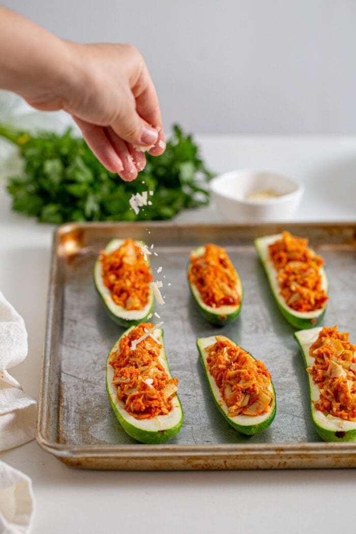 Hand sprinkling cheese over stuffed zucchini boats on a baking tray.